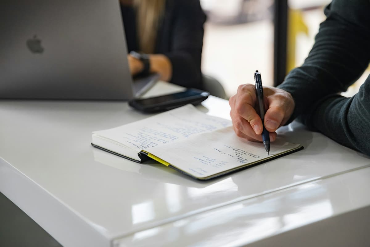Hand schreibt Gesprächsnotizen in ein Notizbuch, daneben Laptop und Smartphone auf einem weißen Tisch